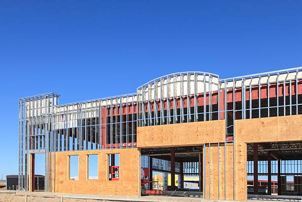 "A new commercial building under construction showing the street front view of the building, metal framing, plywood, door and window openings with blue sky in the background."